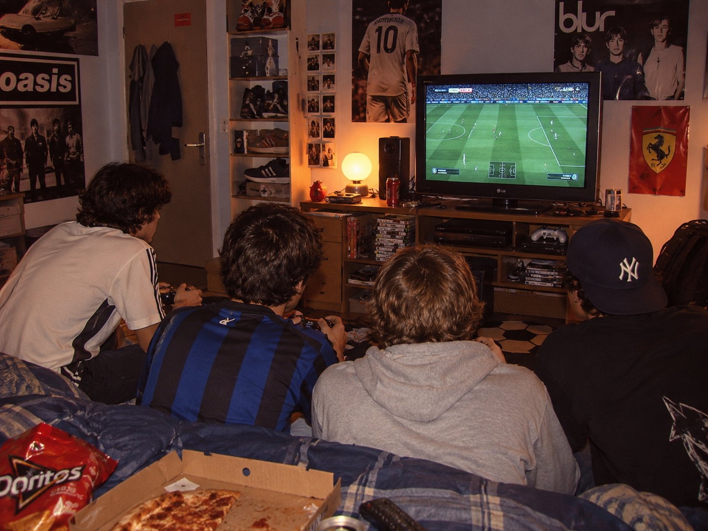 Cuatro amigos de espaldas jugando fútbol en la consola, con pizza y pósters en el cuarto